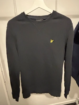 Mörkblå tröja från Lyle & Scott - Snygg svart långärmad tröja från Lyle & Scott med rund halsringning och den klassiska gula loggan broderad på bröstet. Enkel och stilren design som passar perfekt till jeans 