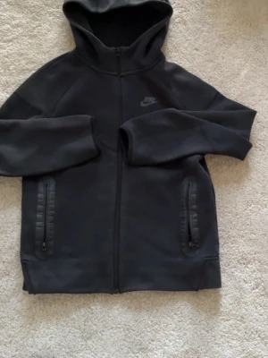 Nike tech hoddie - Säljer en svart hoodie från Nike med dragkedja framtill och två fickor med dragkedja. Klassisk Nike-logga på bröstet och huva. Tillverkad i mjukt material, perfekt för en avslappnad streetwear-look.