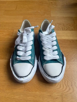 Maison Mihara Green - Snygga gröna sneakers med vit tå och sula, samt breda vita skosnören. Skorna har klassisk låg profil och kontrastsömmar. Ovandelen är i tyg och sulan i gummi, perfekt för en avslappnad streetstil.