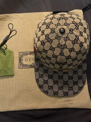 Grå  keps från Gucci med GG-mönster - Säljer en grå keps från Gucci med det klassiska GG-monogrammet över hela kepsen. Kepsen har en rak skärm och svarta detaljer på toppen. Snygg accessoar för dig som gillar lyxiga märken och streetstyle.