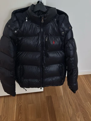 Ralph Lauren mörkblå dunjacka - Snygg mörkblå dunjacka från Ralph Lauren med glansig finish och klassisk puffer-design. Jackan har hög krage, dragkedja framtill, flera fickor och den ikoniska röda Polo-loggan på bröstet. Perfekt för kalla vinterdagar och riktigt stilren.