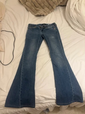 Blåa True Religion flare jeans - Säljer ett par blåa jeans från True Religion, modell Joey Low Rise Flare. Jeansen har låg midja och riktigt utsvängda ben, perfekt för dig som gillar retrovibbar. Klassisk denim med slitningar och kontrastsömmar. Snygga till sneakers eller boots!