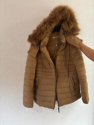 Beige pufferjacka med fuskpäls från MNG - Säljer en beige pufferjacka från MNG med avtagbar huva och fuskpäls. Jackan har dragkedja framtill, två fickor med knappar och quiltad design. Perfekt för kalla dagar och riktigt snygg till alla outfits.