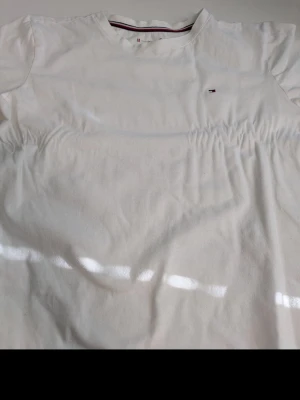 Vit t-shirt från Tommy Hilfiger - Klassisk vit t-shirt från Tommy Hilfiger med liten broderad logga på bröstet. T-shirten har rund halsringning och korta ärmar. Tillverkad i mjuk bomull för en skön känsla. Perfekt till jeans eller shorts för en clean look.