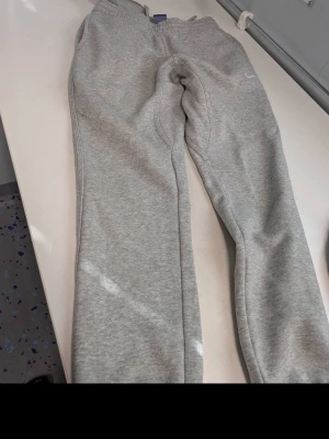 Gråa mjukisbyxor från Nike - Säljer ett par klassiska gråa mjukisbyxor från Nike med resår i midjan och mjukt fleecefoder. Byxorna har en enkel och avslappnad passform, perfekta för chill dagar. Diskret Nike-logga på benet och dragsko i midjan för bästa komfort.