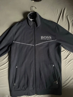 Svart zip hoodie från Hugo Boss - Snygg svart hoodie från Hugo Boss med vit logga på bröstet och vita detaljer längs bröstet. Dragkedja framtill och två fickor med dragkedja. Hög krage och stilren design, perfekt för en casual look.