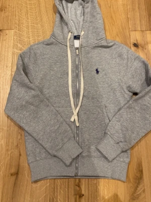 Grå zip hoodie från Polo Ralph Lauren - Snygg grå hoodie med dragkedja från Polo Ralph Lauren. Klassisk design med vit dragsko i huvan och mörkblå logga broderad på bröstet. Tillverkad i mjukt bomullsmaterial och har ribbade muddar vid ärmar och nederkant. Perfekt för en chill och stilren look.