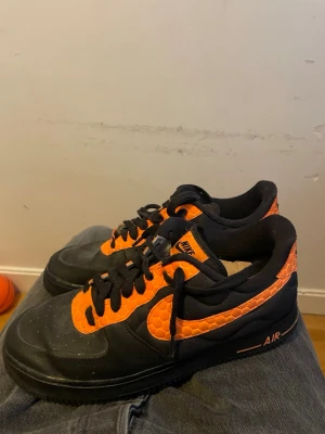 Nike Air Force 1 svart och orange - Nike Air Force 1 sneakers i svart med orange detaljer och ormskinnsmönstrad textur på swoosh och häl. Klassisk låg modell med svart sula och snörning. Perfekt för dig som vill sticka ut med en unik färgkombo och cool design.(Står RIO i insidan av båda tungorna)