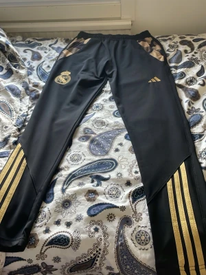 Svarta Real Madrid träningsbyxor Adidas - Snygga svarta träningsbyxor från Adidas med Real Madrid-logga i guld på ena benet och Adidas-logga i guld på andra. Byxorna har guldfärgade detaljer och ränder längs benen samt elastisk midja. Perfekta för fotbollsträning eller chill.