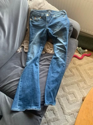 True Religion Joey Low Rise Flare Jeans - Säljer ett par blå True Religion Joey jeans med låg midja och utsvängda ben. Klassisk femficksmodell med snygga kontrastsömmar och detaljerade nitar. Tillverkade i slitstarkt bomullsjeansmaterial. Perfekta för dig som gillar retrovibbar och statement-jeans.