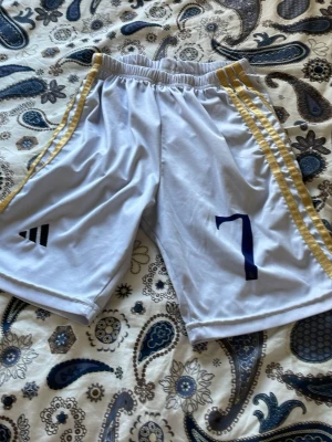 Vita Adidas shorts med gula ränder - Snygga vita sportshorts från Adidas med gula ränder längs sidorna och nummer 7 tryckt på ena benet. Shortsen har elastisk midja och är tillverkade i ett lätt och ventilerande material, perfekt för träning eller match.