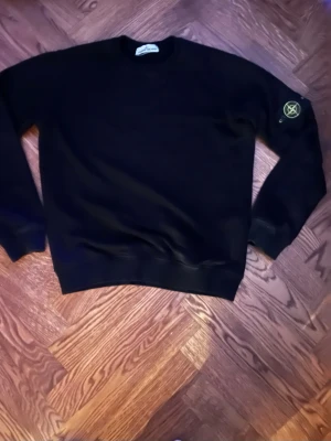 Svart sweatshirt från Stone Island - Snygg svart sweatshirt från Stone Island med klassisk rund hals och ribbade muddar. Ikonisk logotyp-patch på vänster ärm. Perfekt för en stilren och avslappnad look. Tillverkad i mjukt bomullsmaterial. Märket på armen ser bättre ut i verkligheten än på kameran