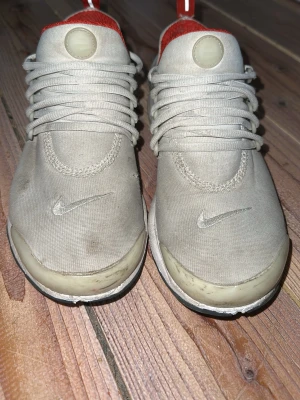 Nike Presto beige och röda sneakers - Nike Presto sneakers i beige med röda detaljer och vit sula. Ovandel i textil och syntet, med röda innerfoder och Nike-logga på sidan och tån. Lågt skaft och snörning framtill. Perfekta för en sportig och avslappnad stil.