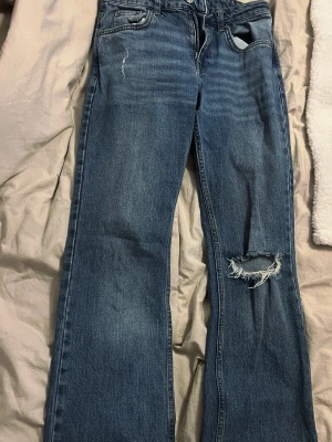 Blå slitna jeans från Perfect Jeans - Ett par blå jeans med raka ben och slitningar, inklusive ett större hål på ena knät. Klassisk femficksmodell med normalhög midja och ljus tvätt. Perfekta för en avslappnad och trendig look.