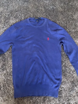Blå stickad tröja från Polo Ralph Lauren - Klassisk blå stickad tröja från Polo Ralph Lauren med rund halsringning och den ikoniska röda logotypen broderad på bröstet. Tröjan har långa ärmar och ribbade muddar vid ärmslut och nederkant. Perfekt för en stilren och avslappnad look. S