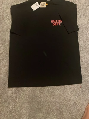 Svart t-shirt från Gallery Dept. - Svart t-shirt från Gallery Dept. med röd logga tryckt på bröstet och stor text på ryggen. Klassisk passform med korta ärmar och rund hals. Tillverkad i mjuk bomull som känns skön mot huden. Perfekt för en stilren och trendig look. Priset kan diskuteras.