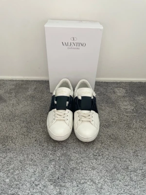 Valentino vita sneakers med svart band - Stilrena vita sneakers från Valentino Garavani med ett brett svart band över vristen. Skorna har klassisk låg profil, vita snören och diskret logga på plösen. Tillverkade i slätt läder med gummisula för extra komfort och grepp.