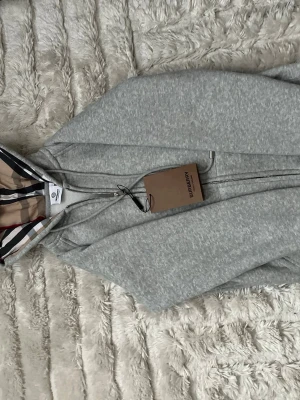 Grå zip hoodie från Burberry - Snygg grå zip hoodie från Burberry med klassiskt rutigt foder i huvan. Dragkedja framtill och justerbar dragsko i huvan. Tillverkad i mjukt bomullsmaterial som känns riktigt skönt mot huden. Perfekt för dig som vill ha en stilren och exklusiv look.