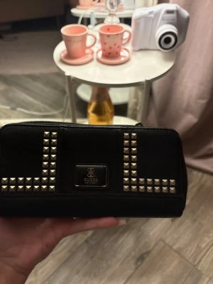 Svart plånbok med nitar från Guess - Snygg svart plånbok från Guess med silverfärgade nitar i rutmönster på framsidan och metallogo. Plånboken har dragkedja runtom, flera fack för kort och sedlar samt ett innerfack med dragkedja. Insidan har ett mönstrat tygfoder i vitt och rött. Helt ny men inget kvitto 