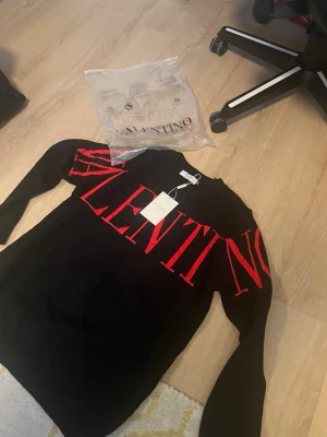 Svart stickad tröja från Valentino - Svart stickad tröja från Valentino med stora röda bokstäver över bröstet och ärmarna. Tröjan har rund halsringning och långa ärmar. Perfekt för dig som gillar statement-plagg och exklusiva märken.