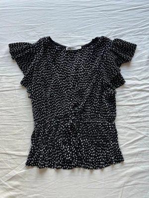 Svartprickig blus från Zara - Söt svart blus från Zara med vita prickar, volangdetaljer vid ärmar och midja samt knytband i midjan. 