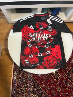 Real Madrid fotboll t shirt - Snygg svart Real Madrid-tröja från Adidas med stora röda rosor och en orm på framsidan. Tryckta vita detaljer, klubbmärke och Adidas-logga på bröstet samt 'Emirates Fly Better' sponsortryck. Korta ärmar och sportig passform, perfekt för fotboll eller streetwear.