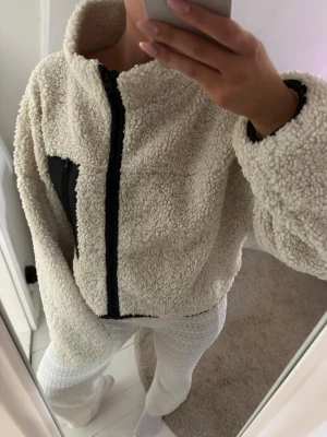Beige teddyjacka med svart detalj - Mysig crémefärgad teddyjacka med dragkedja framtill och svart bröstficka. Jackan har hög krage och oversized passform, perfekt för våren och hösten. Endast använd någon gång i i väldigt fint skick 🌺