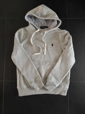 Grå hoodie från Polo Ralph Lauren - Snygg grå hoodie från Polo Ralph Lauren med klassisk huva och vita dragsnören. Broderad marinblå logga på bröstet och stor känguruficka framtill. Tillverkad i mjuk bomullsblandning för skön känsla och avslappnad stil. Passar S/XS