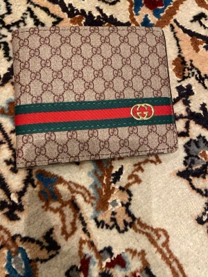  plånbok med GG-mönster - Snygg plånbok från  med klassiskt GG-mönster i beige och brunt. Plånboken har en grön och röd rand med guldfärgad GG-logga på framsidan. Perfekt accessoar för dig som gillar lyxiga detaljer och stilren design.