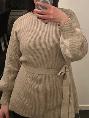 Beige stickad tröja med knytskärp - Snygg beige stickad tröja med ribbad struktur och matchande knytskärp i midjan. Tröjan har rund halsringning och långa ärmar med avslappnad passform. Perfekt för lager på lager och enkel att styla till olika outfits.