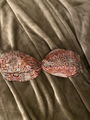 Mönstrad bandeau bikiniöverdel - Säljer en bandeau bikiniöverdel med färgstarkt mönster i orange, rosa, svart och beige. Toppen har ett snyggt blommigt och geometriskt tryck samt silverspänne bak. Perfekt för dig som gillar boho-stil och vill ha något unikt på stranden.