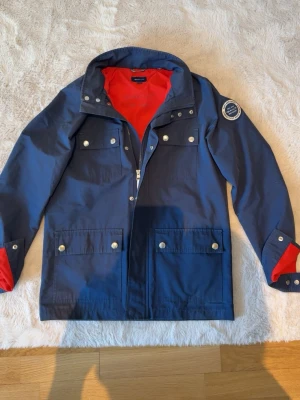 Marinblå jacka med röd insida - Stilren marinblå jacka från GANT med röd insida, fyra fickor framtill med silverfärgade knappar och patch på ena ärmen. Jackan har hög krage och detaljer i rött vid ärmslut och insida. Perfekt för en sportig och höstig look passar för xs 
