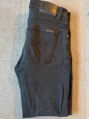 Svarta jeans från Nudie Jeans - Snygga svarta jeans från Nudie Jeans med klassisk femficksdesign och diskret logga på bakfickan. Jeansen har raka ben och är tillverkade i ett mjukt denimtyg med stretch för extra komfort.