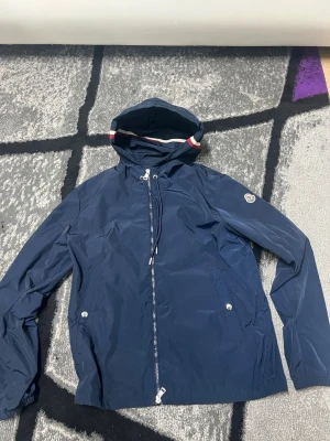 Moncler Windbreaker - Snygg marinblå vindjacka från Moncler med huva och dragkedja framtill. Jackan har två fickor med tryckknappar och en vit-röd-blå rand på huvan. Perfekt för blåsiga dagar och enkel att matcha med olika outfits.