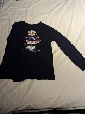 Mörkblå långärmad Polo Bear t-shirt - Snygg mörkblå långärmad t-shirt från Ralph Lauren med ikoniska Polo Bear-trycket på bröstet. Björnen har på sig en USA-tröja och håller en hockeyklubba. Perfekt för dig som gillar sportig och klassisk stil.