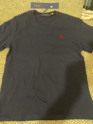 Marinblå t-shirt från Polo Ralph Lauren - Klassisk marinblå t-shirt från Polo Ralph Lauren med rund hals och det ikoniska röda broderade logot på bröstet. Tillverkad i mjuk bomull för en skön känsla och perfekt passform. Enkel och stilren design som funkar till allt.