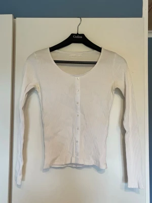 Vit ribbad tröja med knappar - Säljer en vit ribbad tröja med långa ärmar och rund halsringning. Tröjan har knäppning framtill med små vita knappar och är tillverkad i ett stretchigt material som sitter snyggt på kroppen. Perfekt att styla över ett linne eller t-shirt.
