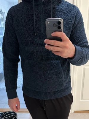 Mörkblå teddy hoodie med huva - Mysig mörkblå hoodie från Peak Performance i teddy-material. Har stor magficka, broderad logga på bröstet och justerbar huva med dragsko. Perfekt för dig som gillar en avslappnad och trendig stil.