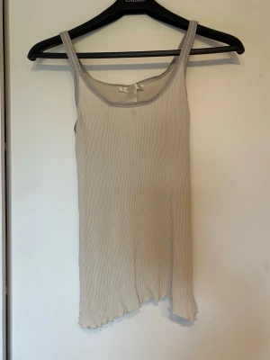 Ribbad beige linnetopp First Class - Säljer en ribbad beige linnetopp från First Class. Toppen har smala axelband och ärmlös design med stretchigt material och asymmetrisk nederkant. Perfekt för en enkel och stilren look.