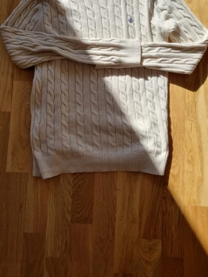 Beige kabelstickad tröja Morris - Kabelstickad tröja i beige från Morris, tillverkad i 100% pima bomull. Klassisk modell med ribbade muddar och diskret logga broderad på bröstet. Perfekt för dig som gillar stilrena och tidlösa plagg.