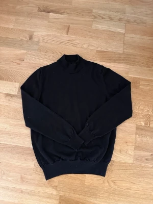 Svart polotröja från J.Lindeberg - Klassisk svart stickad polotröja från J.Lindeberg. Tröjan har lång ärm, ribbade muddar och hög krage. Perfekt basplagg i garderoben och enkel att matcha med olika stilar. Materialet känns mjukt och följsamt. Storlek L men passar nog S/M också då den är liten i storlek och sitter rätt tajt.