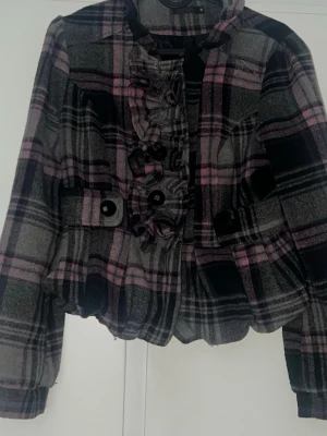 🖤 Rutig jacka med puffig passform – stl M (passar även S) - Snygg rutig jacka i grått, svart och rosa med volangdetaljer längs knappraden. Jackan har stora svarta knappar framtill och på fickorna, samt en croppad passform. Perfekt för dig som vill sticka ut med en unik stil.🍂🍁. Obs: Två knappar framtill är öppna/behöver sys fast igen (enkelt fix 🪡) ( kan gärna fixa de ) jackan passar även de som har storleken S perfekt ” det står M i lappen men den är inte så 