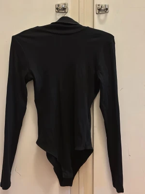 Svart långärmad body från H&M - Snygg svart body från H&M med lång ärm och hög hals. Tillverkad i ett stretchigt och mjukt material som sitter tight mot kroppen. Perfekt att styla med jeans eller kjol för en clean look.