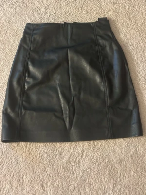 Svart kort kjol i skinnimitation - Snygg svart kort kjol i skinnimitation med dragkedja bak. Kjolen har en stilren och enkel design som passar till många olika looks. Perfekt för dig som gillar en edgy och trendig stil.