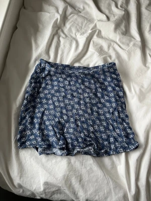 Blå mönstrad kortkjol från H&M - Säljer en blå kortkjol från H&M med vitt geometriskt mönster. Kjolen är gjord i mjuk bomull och har en avslappnad passform. Perfekt för dig som gillar en enkel men snygg stil.