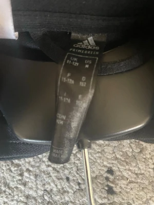 Svart Adidas träningströja herr - Svart långärmad träningströja från Adidas med hög krage och vita detaljer längs ärmarna. Tillverkad i Primegreen-material som är återvunnet och funktionellt. Perfekt för dig som gillar sportig stil och vill ha något bekvämt till träningen.