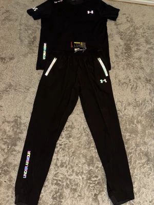 Svart träningsset från Under Armour - Snyggt svart träningsset från Under Armour med t-shirt och tights. T-shirten har färgglada logotyper och korta ärmar. Tightsen har dragkedja och logga på benet. Perfekt för gymmet eller löpning, tillverkad i ett lätt och stretchigt material.