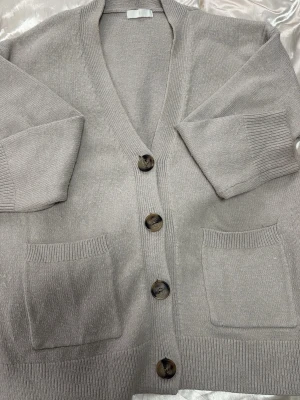 Beige kofta med stora knappar - Mysig beige kofta med v-ringning och fyra stora bruna knappar framtill. Koftan har två rymliga fickor och ribbade muddar vid ärmslut och nederkant. Perfekt för lager-på-lager och enkel att matcha med olika outfits.