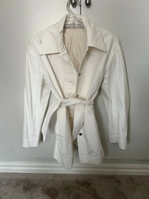 Vit trenchcoat från Filippa K - Stilren vit trenchcoat från Filippa K med klassisk krage, knäppning framtill och matchande midjebälte. Tillverkad i ett kraftigare bomullstyg som ger en snygg och tidlös look. Perfekt för dig som gillar minimalistisk och clean stil.