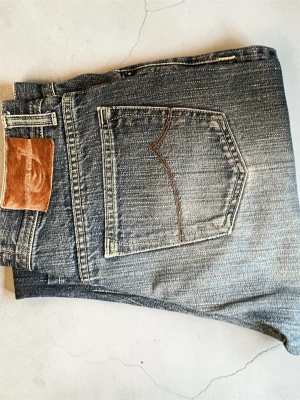 Blå jeans från Jacob Cohën (Nyskick) - Snygga blå jeans från Jacob Cohën med klassisk femficksmodell och kontrastsömmar. Jeansen har en unik patch i brunt läder med hårdetalj och broderad logga bak i midjan. Tillverkade i slitstark denim med tvättad look och raka ben. Jeansen är i nyskick bortsett från en sömm som har gått upp på ena benet längst ner. (Kan gå ner i pris)
